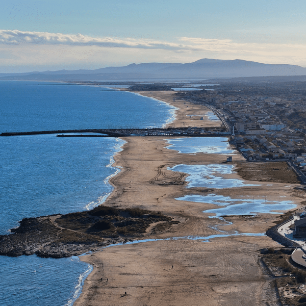 Panorama du Littoral Méditerranéen et ses Plages de Sable Vue aérienne de la côte méditerranéenne, plages de sable, lagunes et montagnes lointaines sous un ciel ensoleillé.