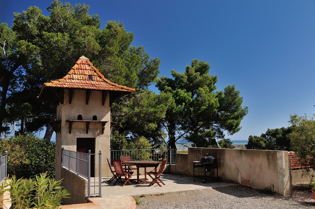 Terrasse ensoleillée avec petite tour rustique, table en bois, barbecue et vue sur mer entre les pins méditerranéens.